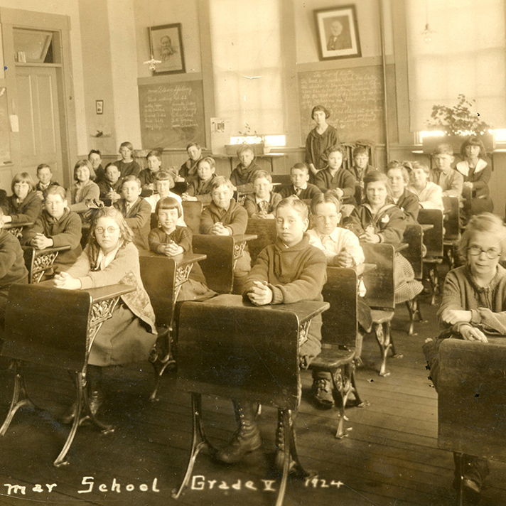 Sepia-toned historical photograph of an early 20th-century classroom filled with young students seated at wooden desks arranged in rows. The children are dressed in period clothing, with many clasping their hands on their desks and facing forward. A teacher stands at the back of the room, and another adult is visible to the side. The classroom features tall windows letting in natural light, framed portraits and writing on the chalkboards, and potted plants on the windowsills. The image is labeled "Grammar School Grade V 1924" at the bottom.
