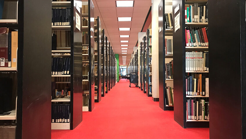 View down a long, symmetrical aisle between tall library bookshelves filled with books. The aisle is carpeted in bright red and extends toward a windowed area in the distance under a ceiling of rectangular lights.