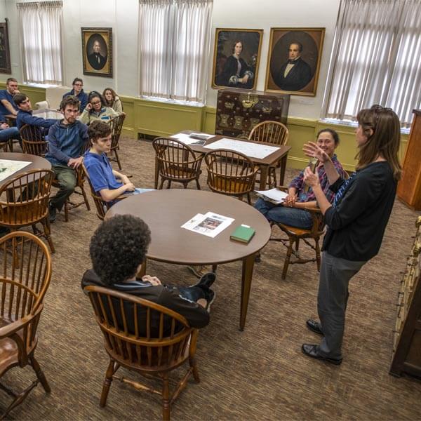 A group of people sit around a wooden table in a well-lit room with portraits hanging on the walls. One person, standing at the front of the group, is speaking and gesturing with their hands, while others listen attentively. The group consists of several individuals of varying ages and genders, with a mix of seated and standing participants. The room has large windows with curtains and is furnished with wooden chairs and tables.