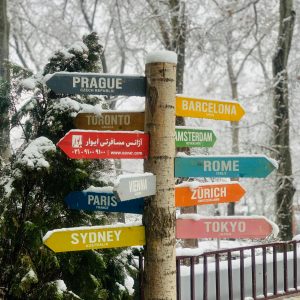 Snow-covered wooden post with directional signs pointing to cities like Prague, Toronto, Tokyo, and Paris.