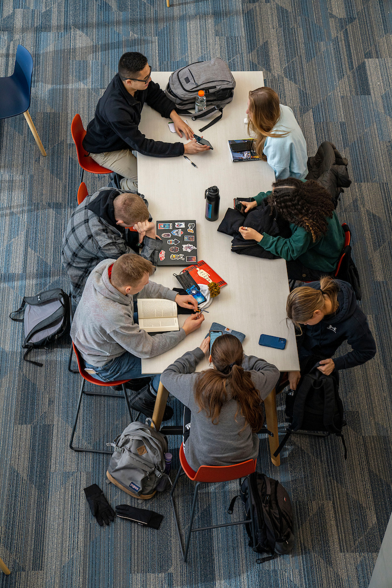 An overhead view of seven students sitting around a rectangular table in a study area with blue carpeted flooring. Some students are using laptops or smartphones, while others are reading or talking. The group appears to be engaged in collaborative work or studying in a casual academic setting.