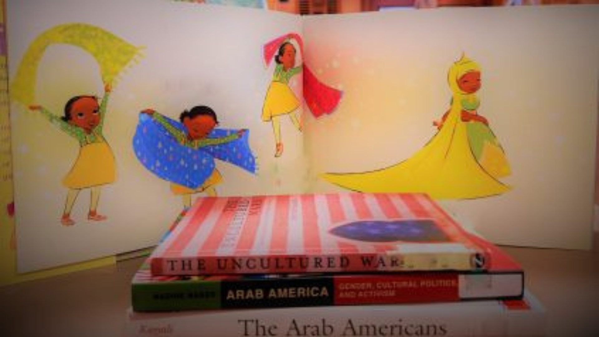 A stack of books on Arab-American heritage and culture, including titles like Arab America, positioned in front of an open children’s book illustrating a young girl wearing a yellow hijab.