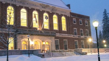 Fogler Library during dusk, with its lights on and surrounded by snow