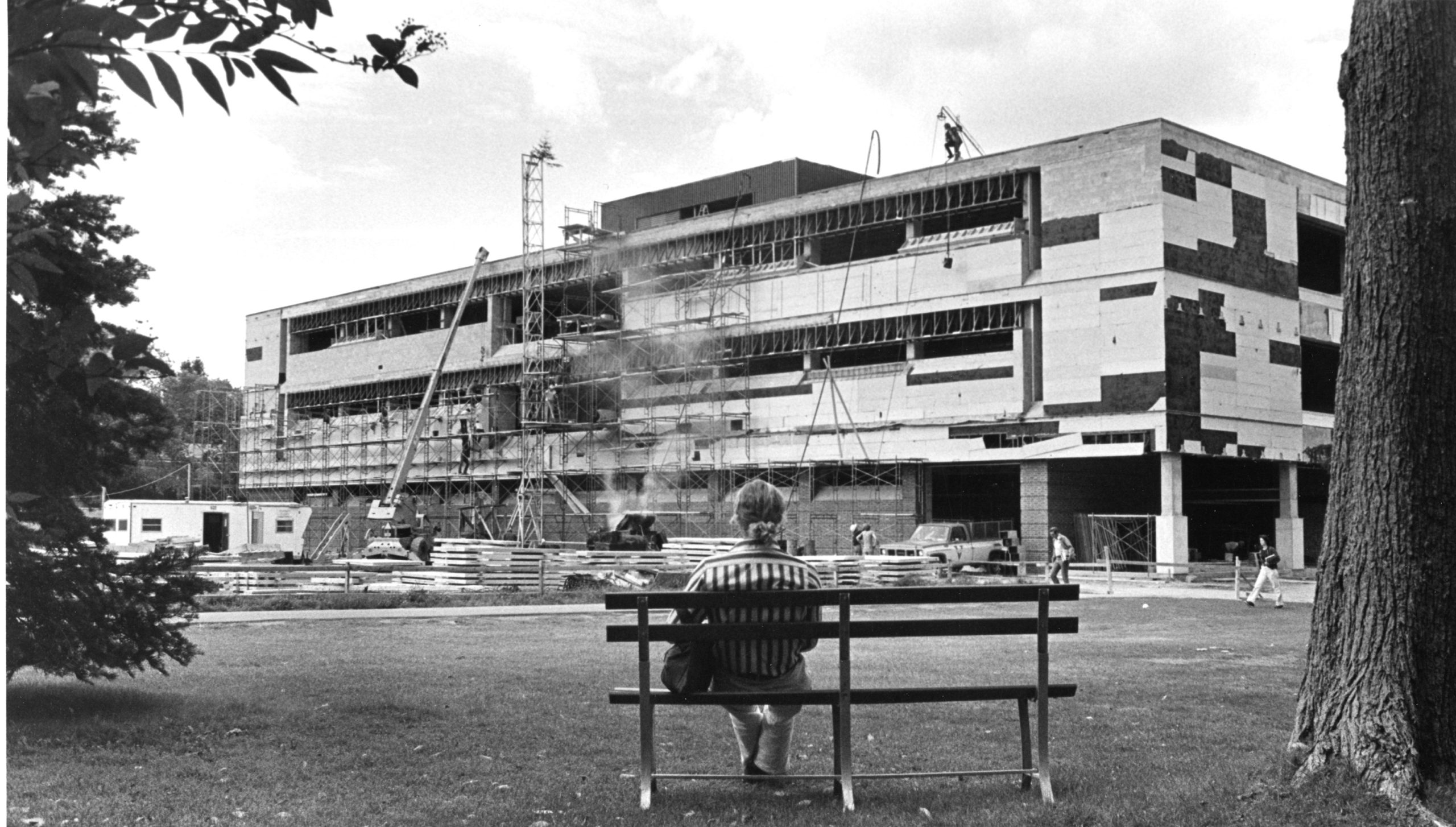 Black-and-white photo of the University of Maine’s Fogler Library under construction. Workers stand on scaffolding and a crane is visible at the site. A student sits on a bench in the foreground, observing the progress. The building's facade is partially complete.