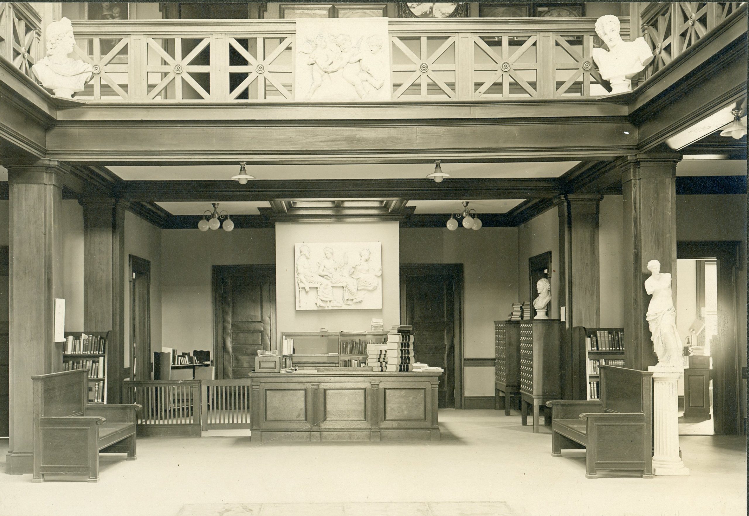 Sepia photo of a formal academic library lobby featuring classical statues, busts, bookshelves, and a wood-paneled information desk. Decorative plaster reliefs adorn the walls above the main desk.