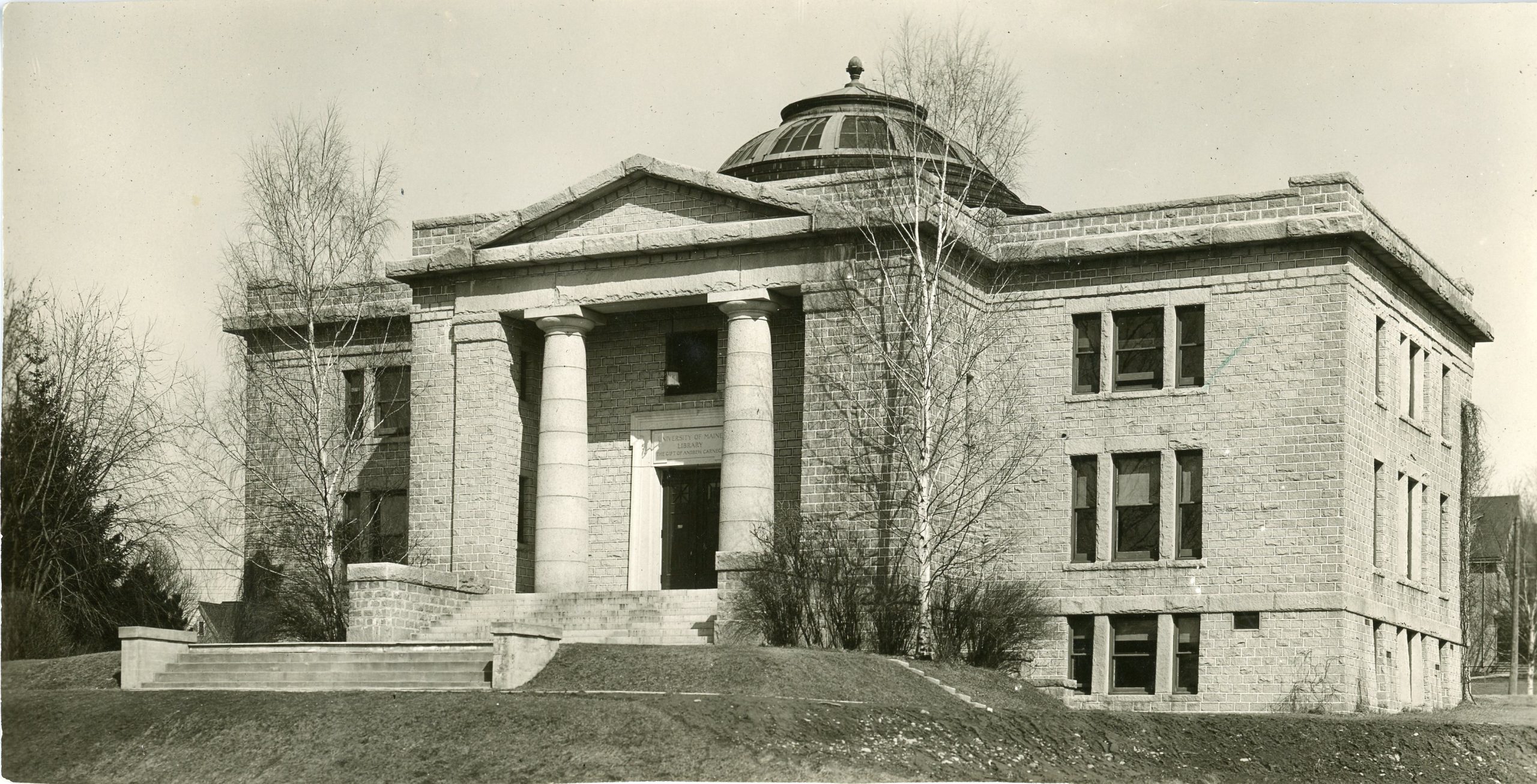 Historic photo of the exterior of the University of Maine’s Carnegie Library. The neoclassical stone structure has large columns flanking the main entrance and a domed roof. Leafless trees frame the building, indicating late fall or early spring.