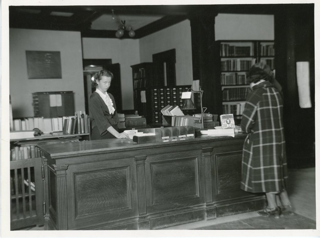 Black-and-white photograph of a female librarian assisting a patron at the circulation desk in the University of Maine’s library. Books, card catalogs, and wooden furniture are visible throughout the room.
