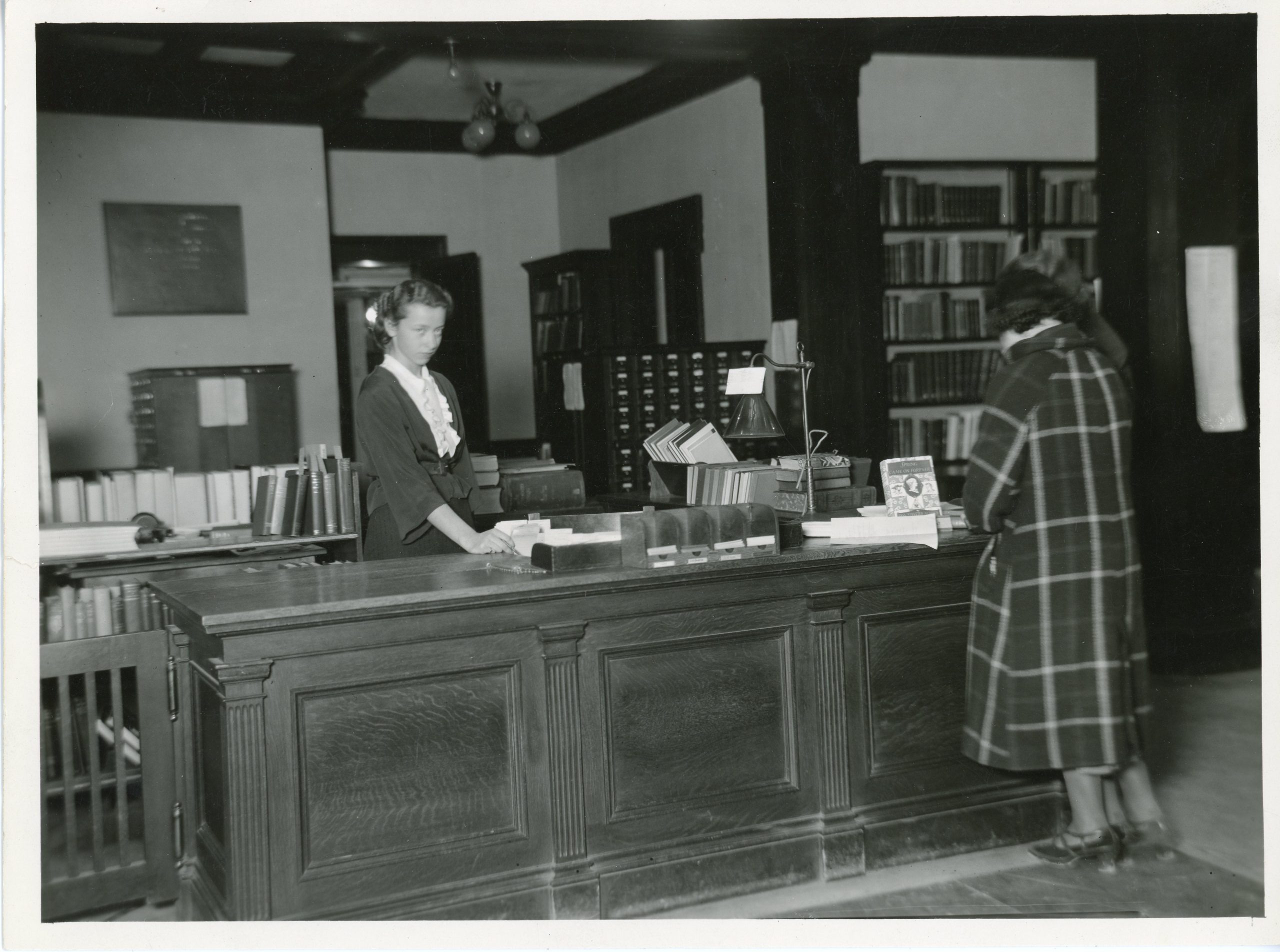 Black-and-white photograph of a female librarian assisting a patron at the circulation desk in the University of Maine’s library. Books, card catalogs, and wooden furniture are visible throughout the room.