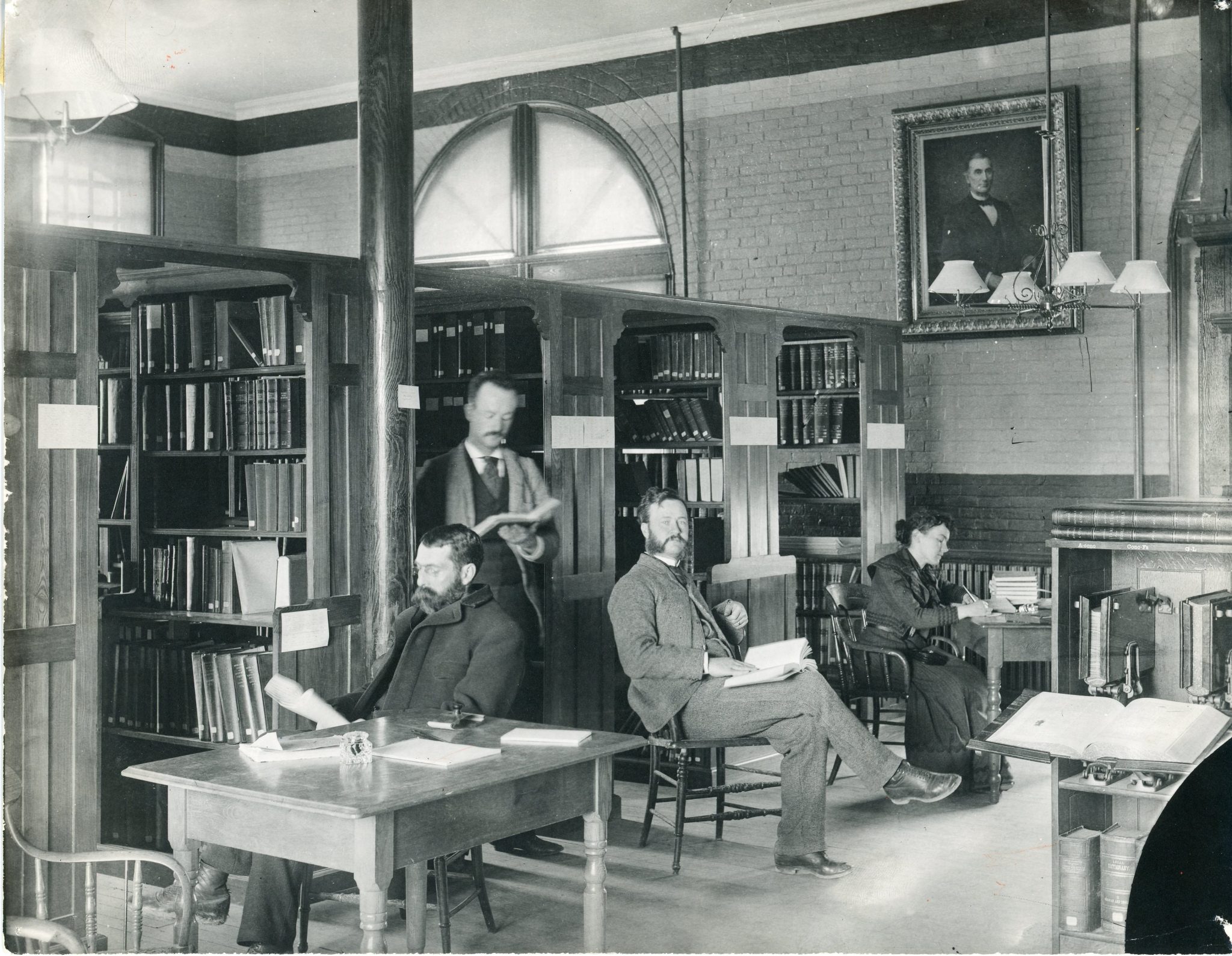 Turn-of-the-century photo showing several men and a woman seated at tables inside a library, reading and working. The room features arched windows, wooden bookshelves, and a portrait above the fireplace.