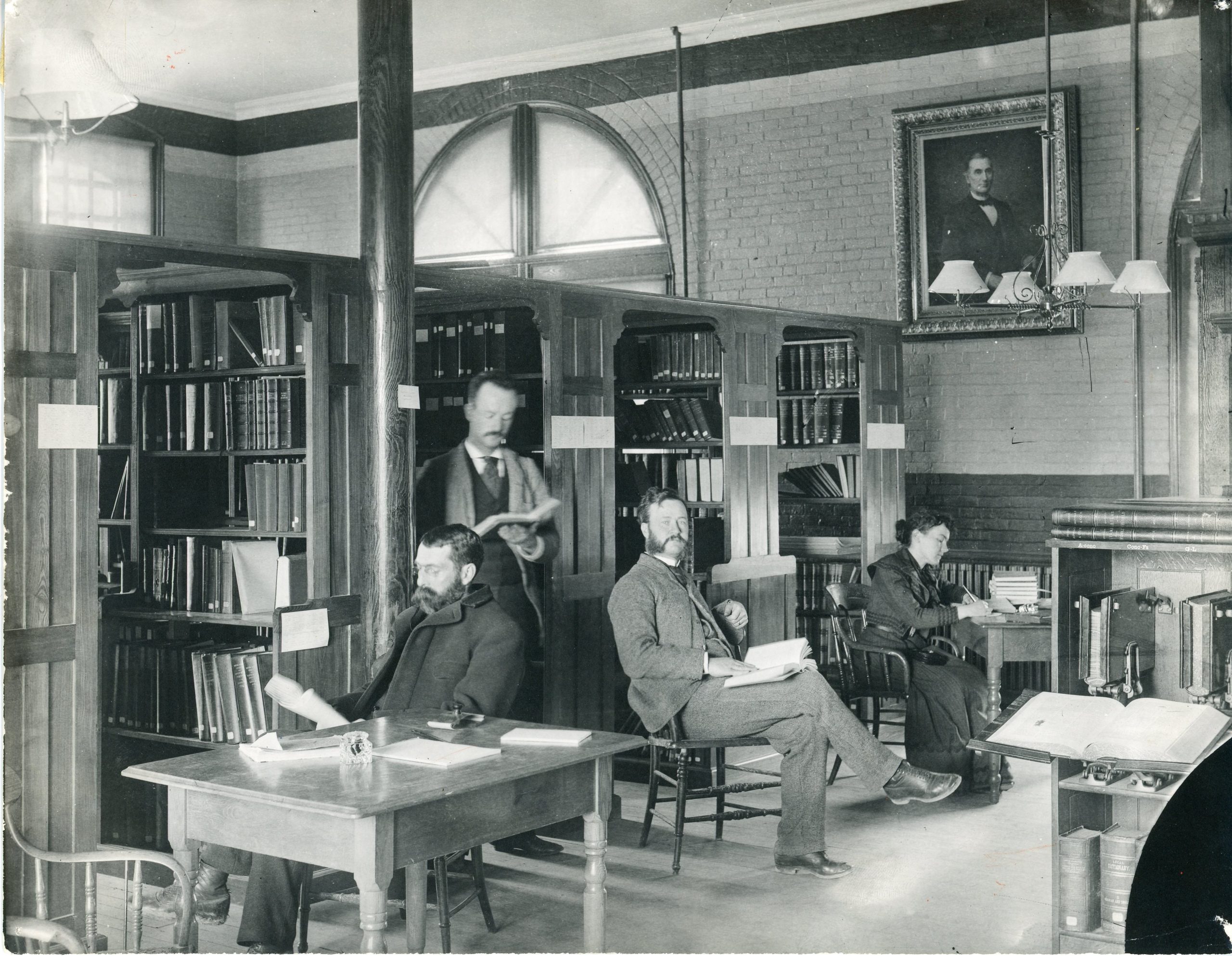 Turn-of-the-century photo showing several men and a woman seated at tables inside a library, reading and working. The room features arched windows, wooden bookshelves, and a portrait above the fireplace.
