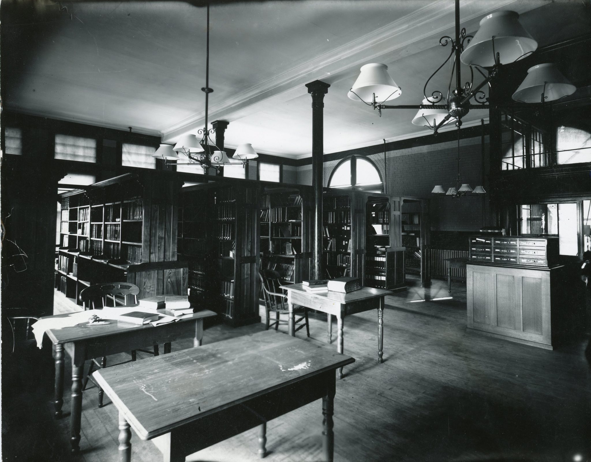 Black-and-white photograph of an early 20th-century academic library reading room with tall bookshelves, wooden tables, spindle-back chairs, and hanging chandeliers. Daylight filters in through high windows, illuminating the dark wood interior. A card catalog is visible on the right.