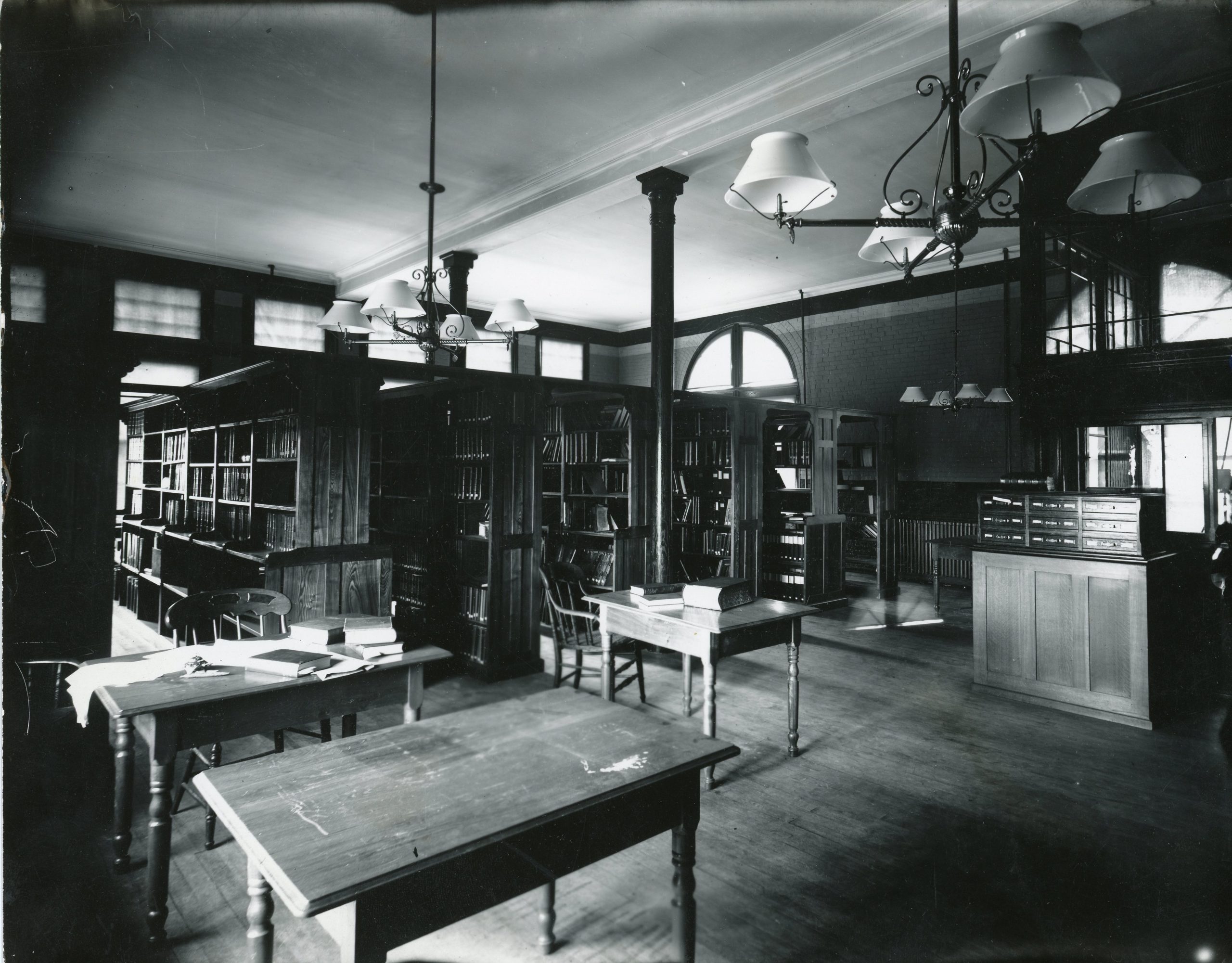 Black-and-white photograph of an early 20th-century academic library reading room with tall bookshelves, wooden tables, spindle-back chairs, and hanging chandeliers. Daylight filters in through high windows, illuminating the dark wood interior. A card catalog is visible on the right.