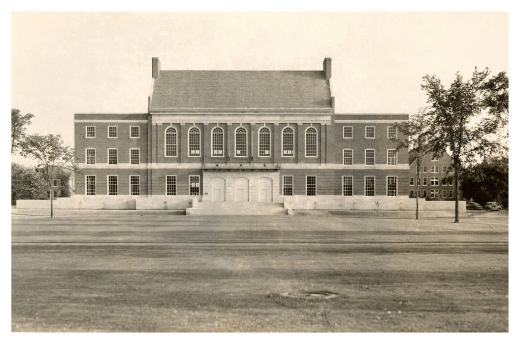 Historic photo of the completed Fogler Library. The symmetrical brick building features large arched windows, tall white doors, and a broad staircase leading up from the lawn. Trees frame the edges of the image.