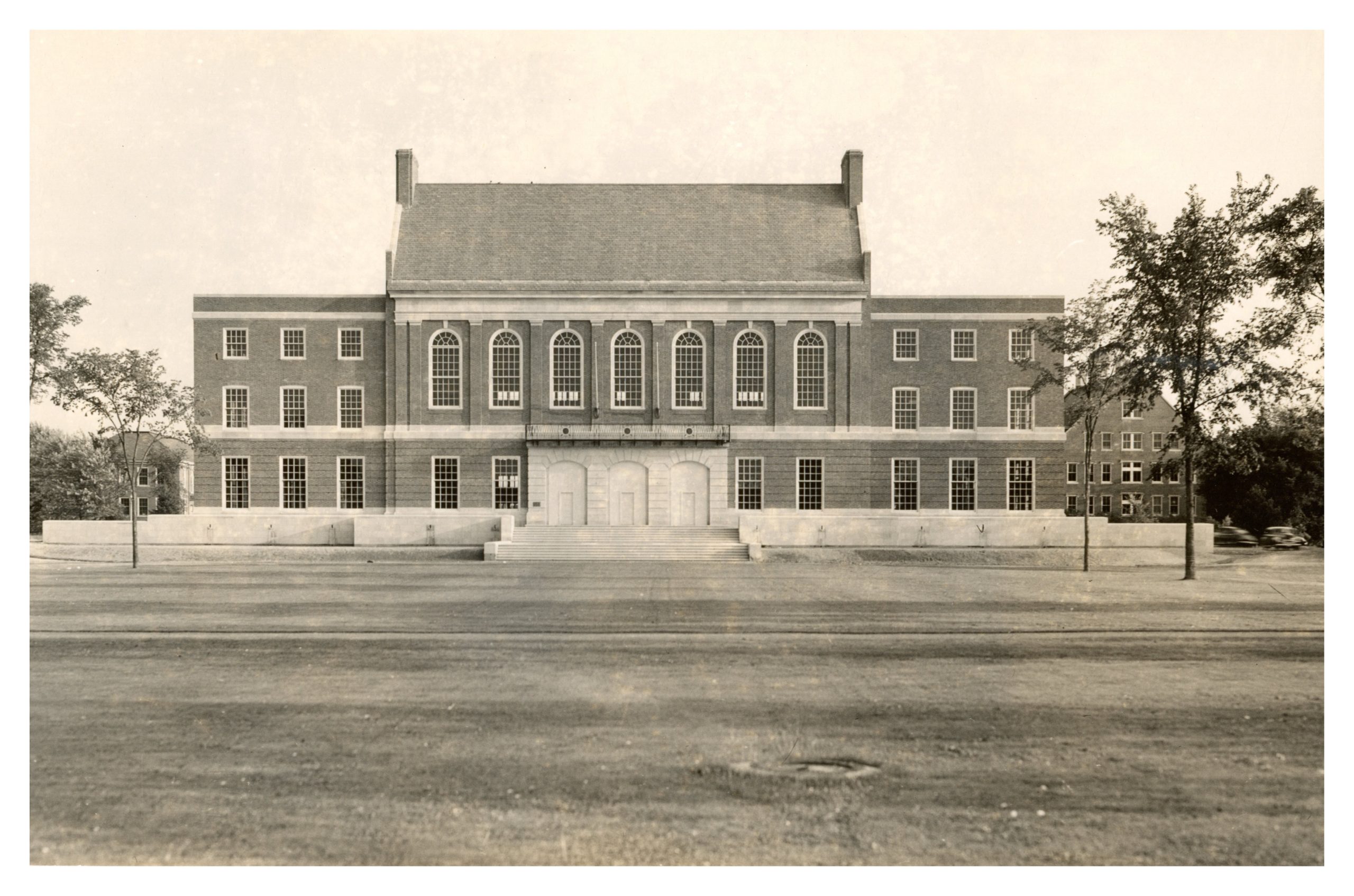 Historic photo of the completed Fogler Library. The symmetrical brick building features large arched windows, tall white doors, and a broad staircase leading up from the lawn. Trees frame the edges of the image.