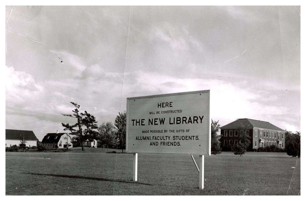 Black-and-white image of a sign on the University of Maine campus that reads “HERE WILL BE CONSTRUCTED THE NEW LIBRARY,” with a note about alumni, student, faculty, and friend support. The existing campus buildings are visible in the background under a partly cloudy sky.